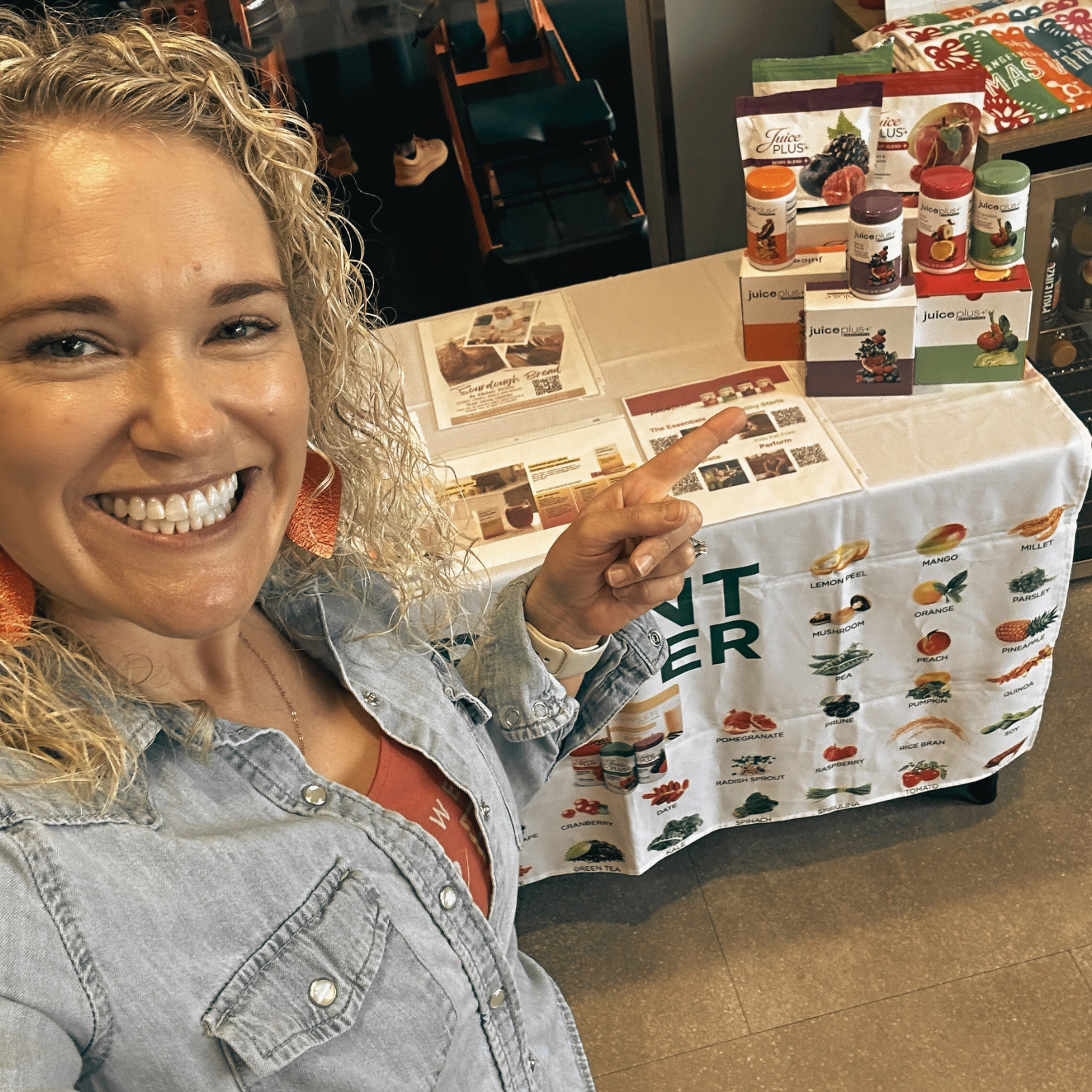 Juice Plus: Bridging the Gap for Optimal Health Rachael DeBoy smiling and pointing to a Juice Plus display table featuring various product blends, emphasizing how Juice Plus helps bridge the gap between the fruits, vegetables, and berries we eat and what our bodies need for optimal health.
