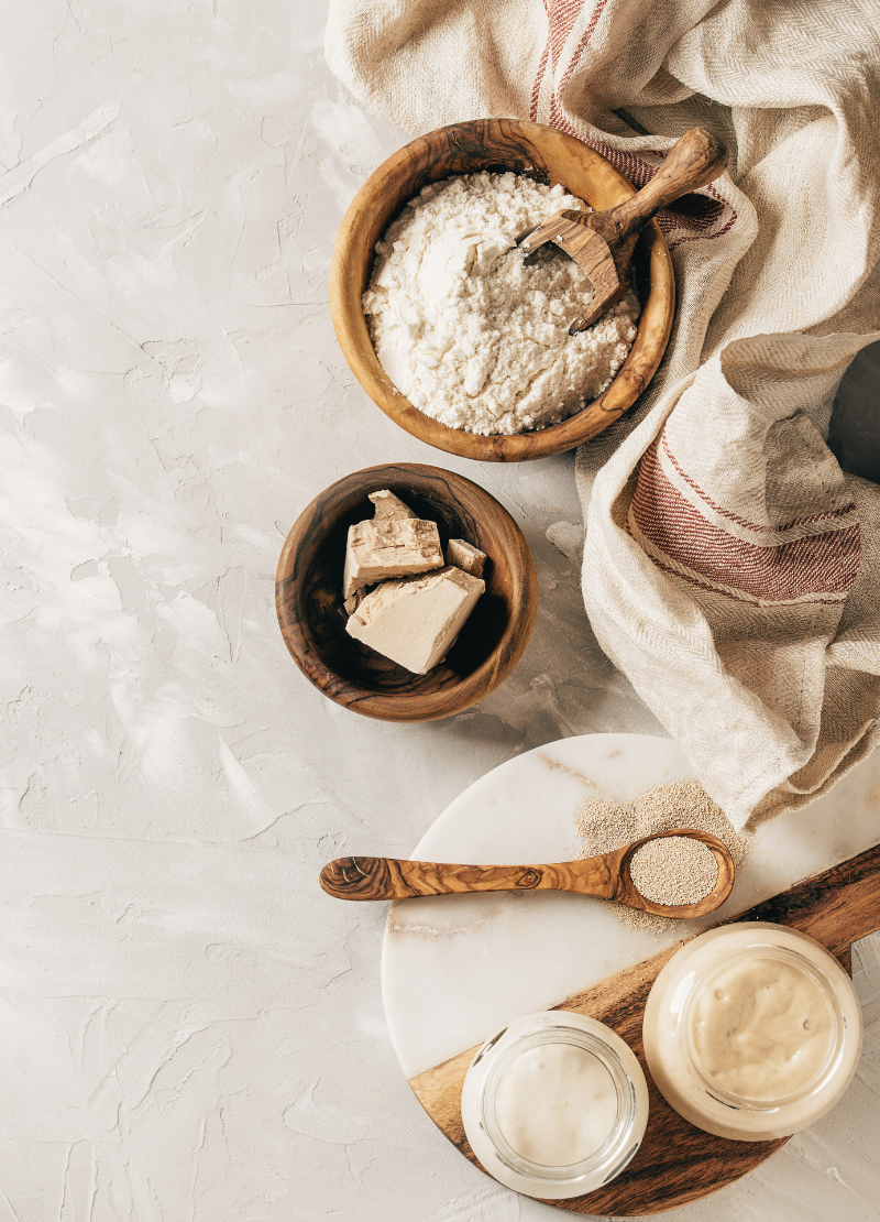 Sourdough: A Healthier and More Delicious Way to Bake Wooden bowls with flour and natural yeast, alongside a glass jar of sourdough starter, representing the essential ingredients for sourdough baking and highlighting its health benefits over conventional bread.
