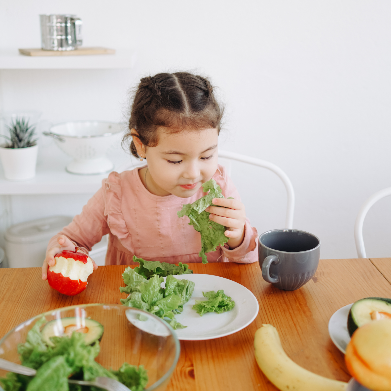Encouraging Kids to Eat More Fruits and Veggies: Simple Tips A young girl sitting at a table, holding an apple and a leaf of lettuce, enjoying healthy food choices. The image illustrates tips for parents on how to get kids to eat more fruits and vegetables.