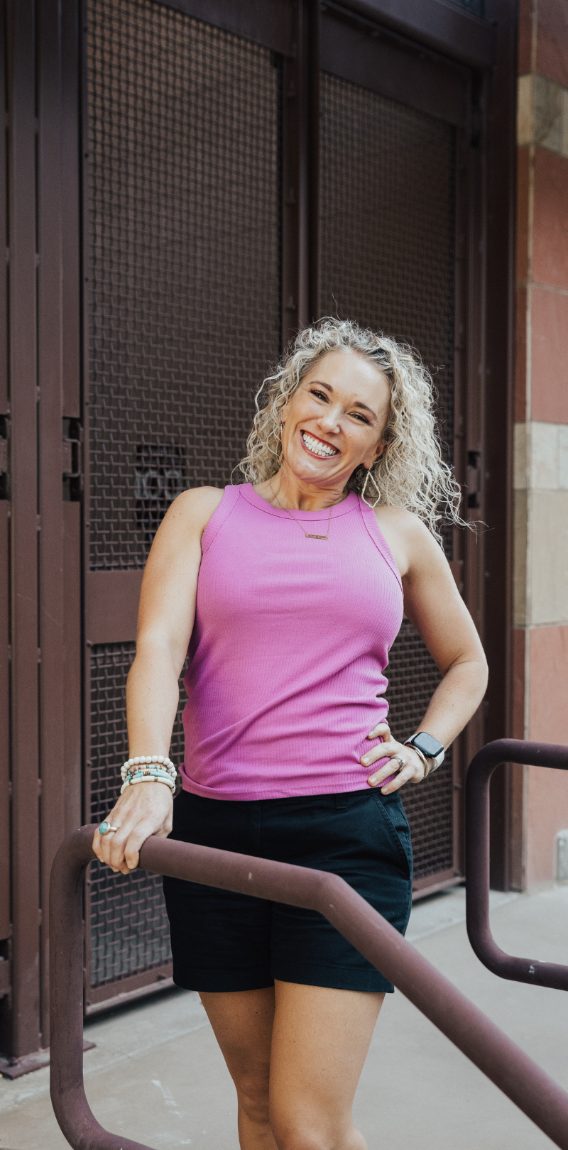 Image of Rachael DeBoy, founder of Wellness with Rachael, smiling and standing confidently against a metal and brick backdrop. She specializes in sourdough mentoring, financial guidance, insurance advocacy, and meal planning, helping people build a solid foundation for health and financial stability.