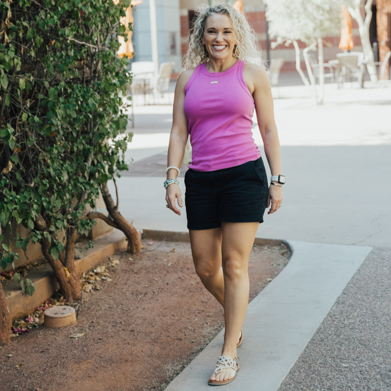 Rachael DeBoy, wellness educator and owner of Wellness with Rachael in San Antonio, Texas, smiling while walking outdoors in a pink top and black shorts.