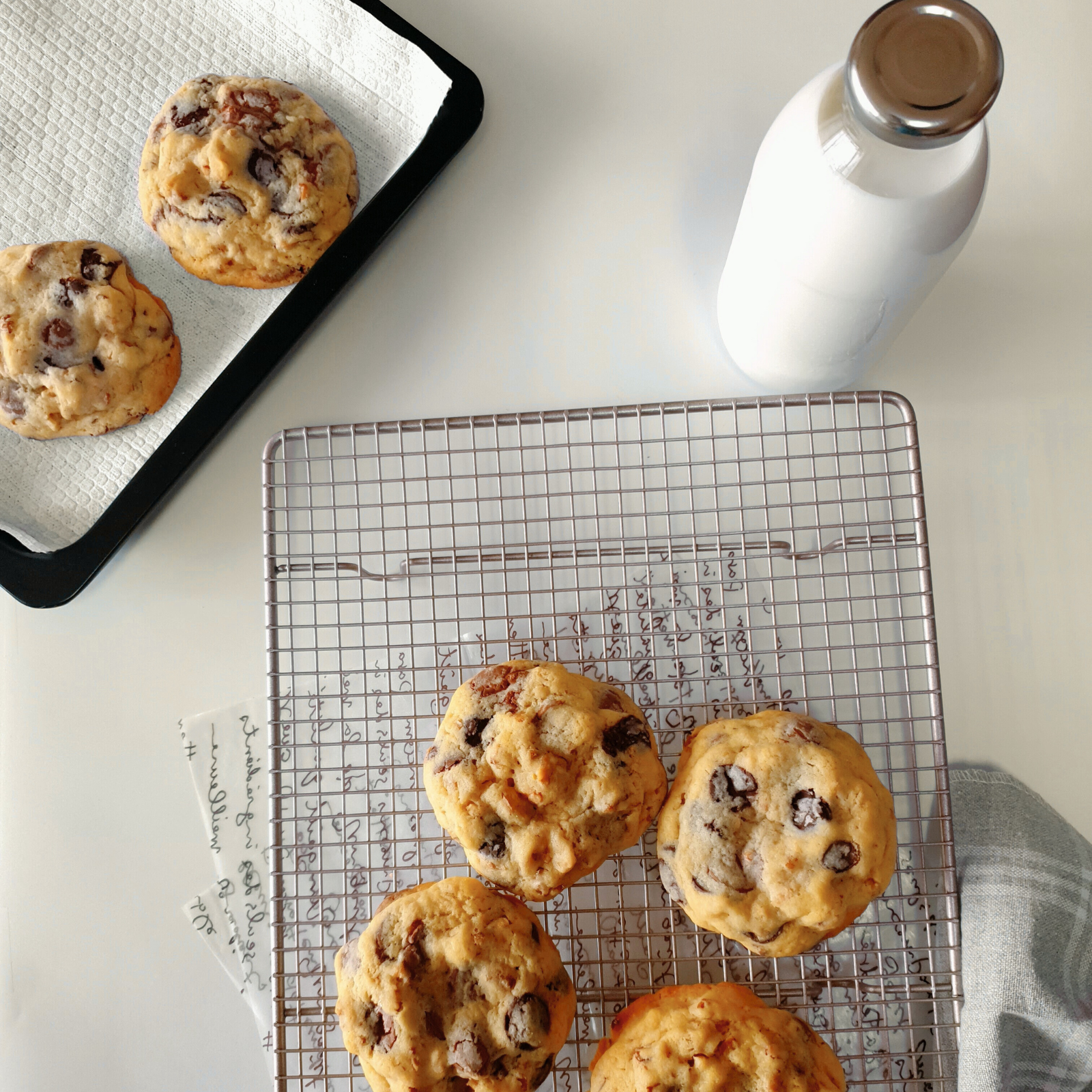 Freshly baked chocolate chip cookies on a wire rack next to a bottle of milk and a baking tray on a white countertop.