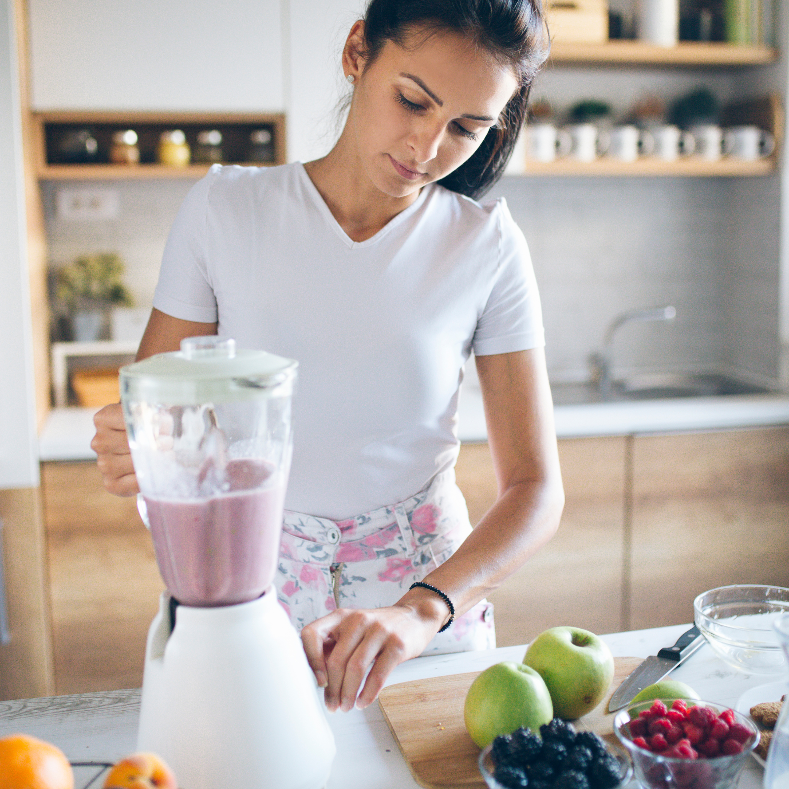 Woman in a white shirt blending a smoothie surrounded by fresh fruit like apples, berries, and oranges