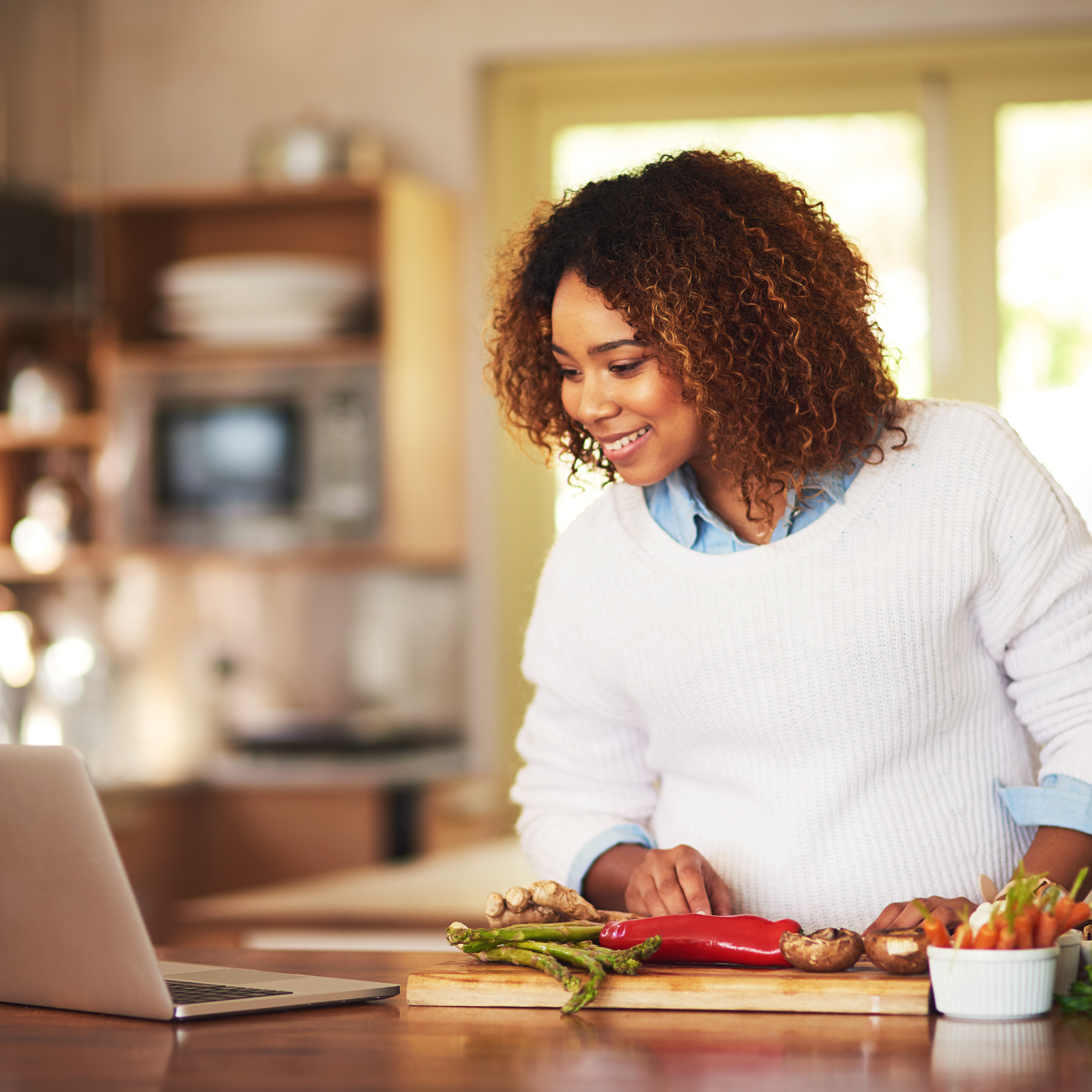 Smiling woman in a kitchen preparing fresh vegetables while watching a laptop, representing easy meal planning and home cooking for families.