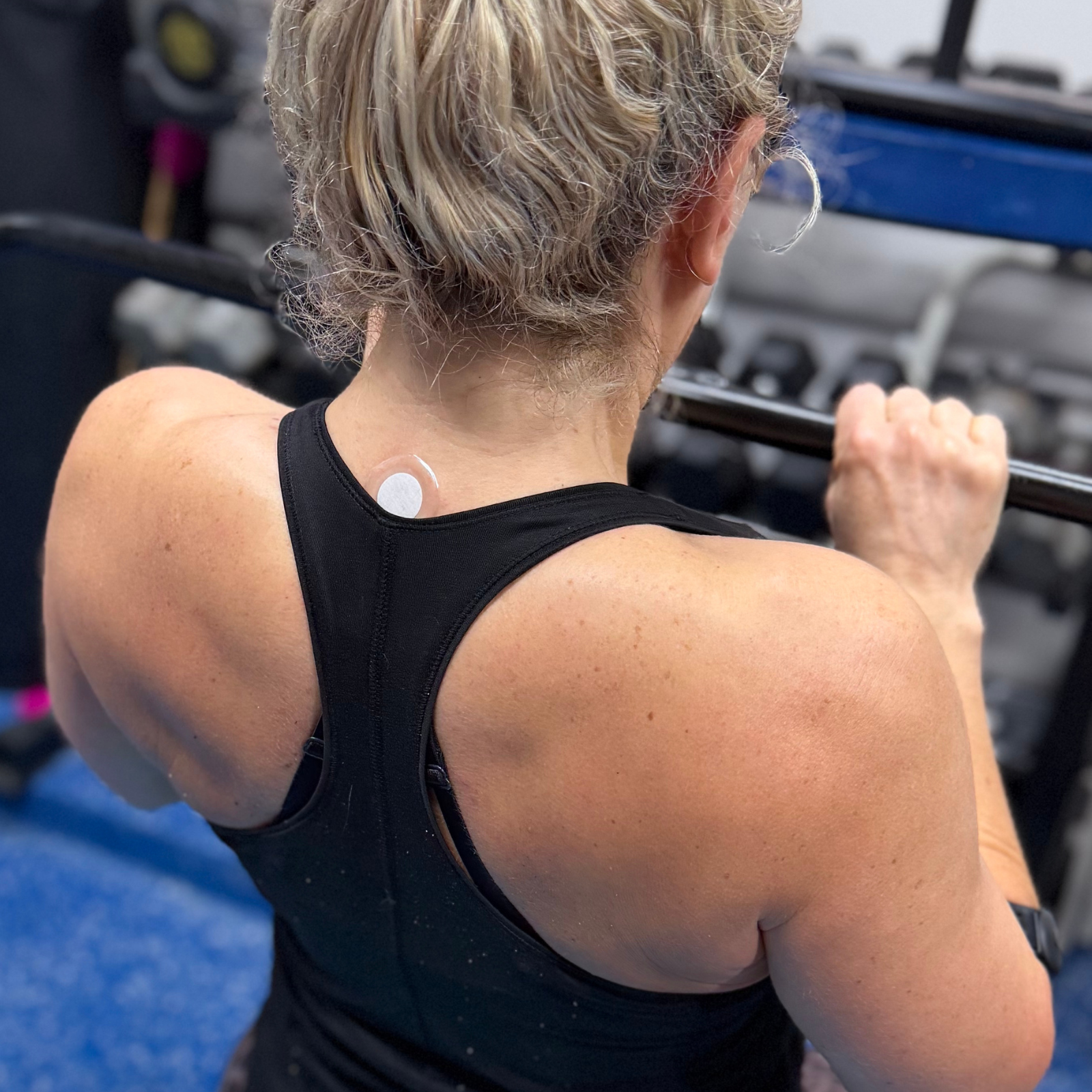 Woman lifting weights in a gym while wearing a LifeWave patch on her neck, shown from behind to highlight patch placement and active lifestyle.