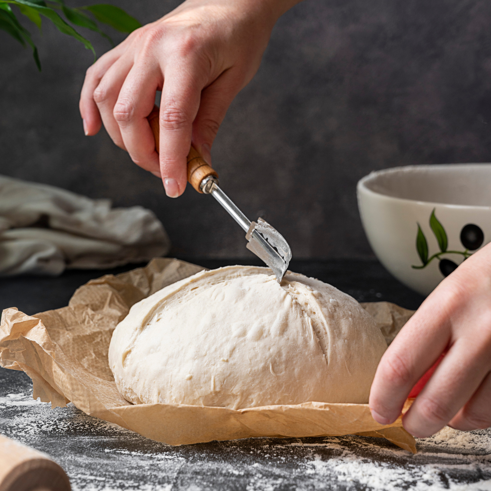 Close-up of hands scoring sourdough bread dough with a lame on parchment paper